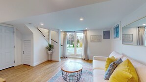 A modern living room with a staircase, a white sofa with yellow cushions, a round coffee table, and a view of the outside.