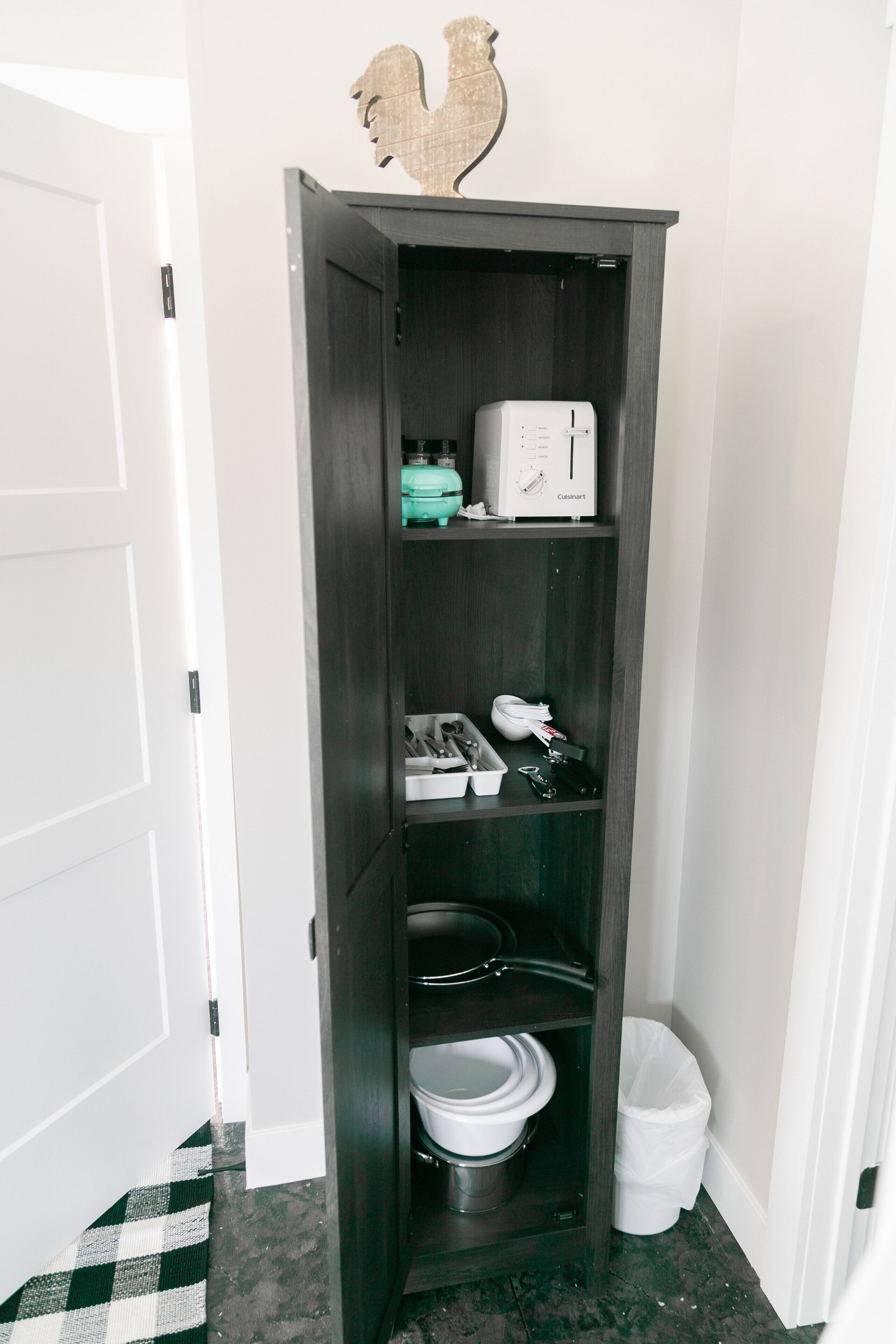 A narrow, dark wooden cabinet with shelves containing a white toaster, a green container, and a white trash bin.