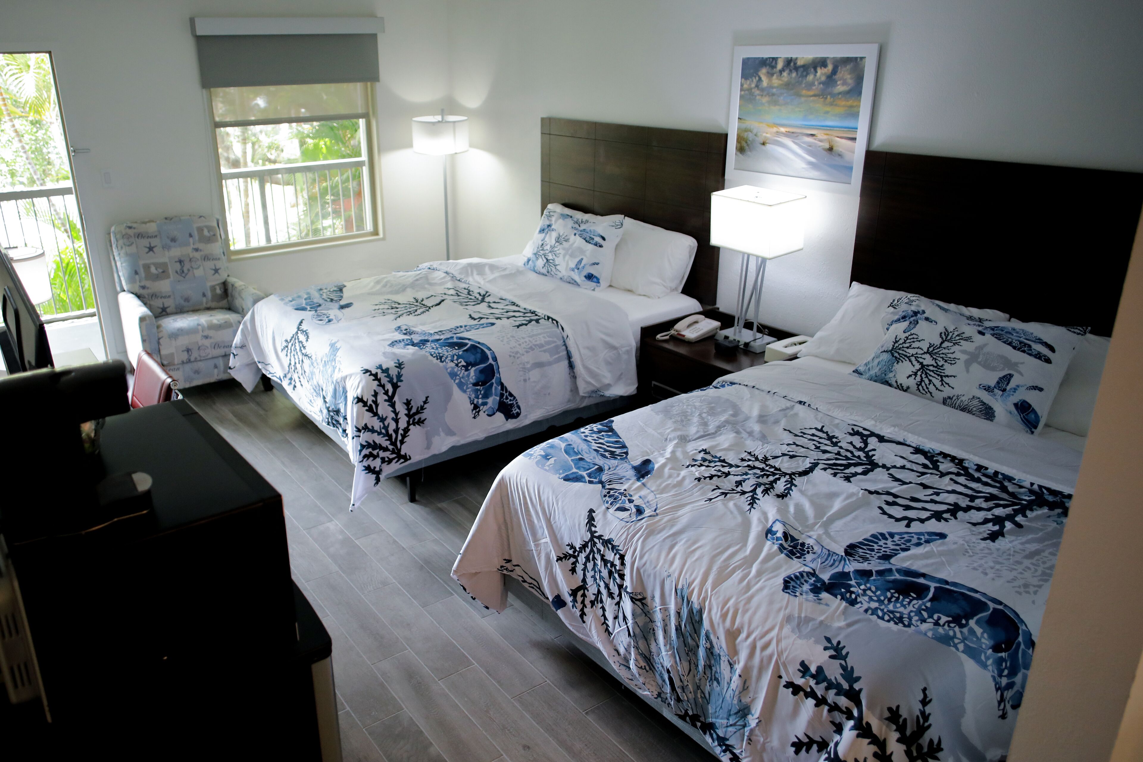 Quarto de hotel com duas camas, uma cadeira, uma escrivaninha e janela com vista para o verde.