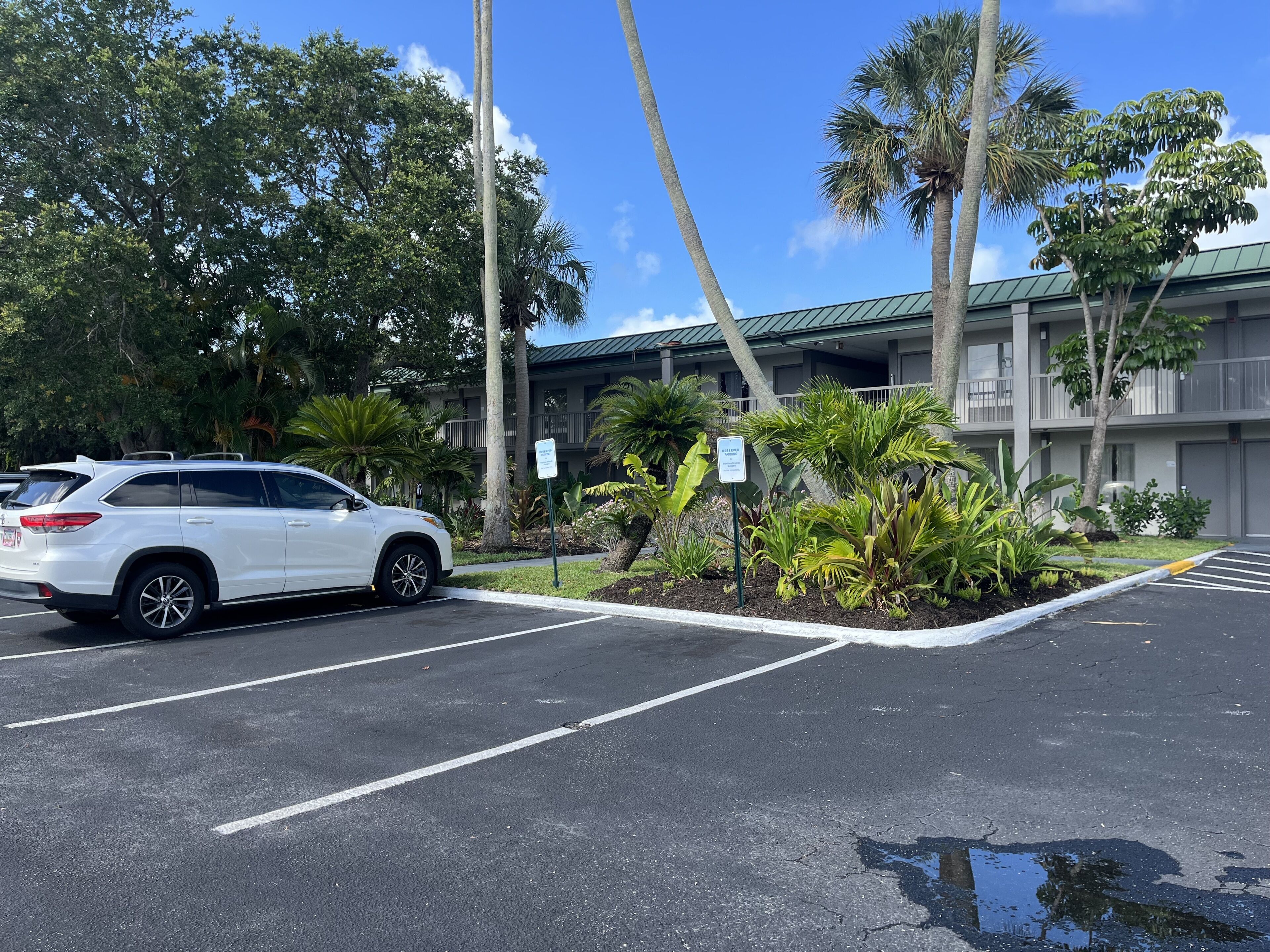 Un SUV blanco estacionado en un estacionamiento, con un edificio de balcones y palmeras al fondo.