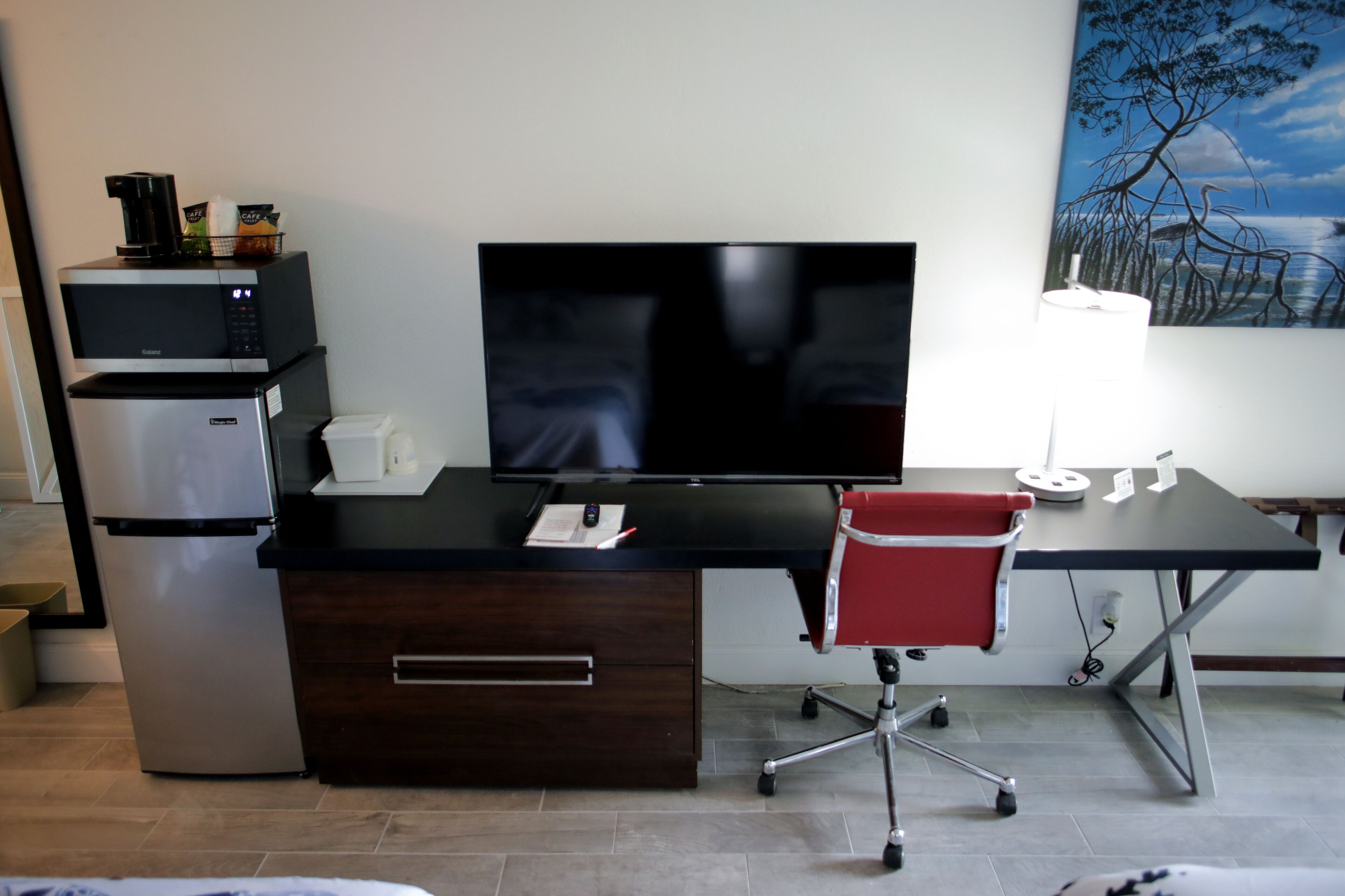 Una camera d'hotel con una scrivania, una TV a schermo piatto, un frigorifero, un forno a microonde, una poltrona da ufficio rossa e un quadro appeso al muro.