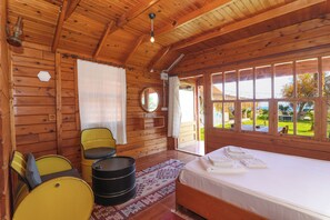 A wooden cabin room with a bed, two chairs, a small table, and a window with a view of the outdoors. - Demre Sahildeki Ev Bungalov (Demre)
