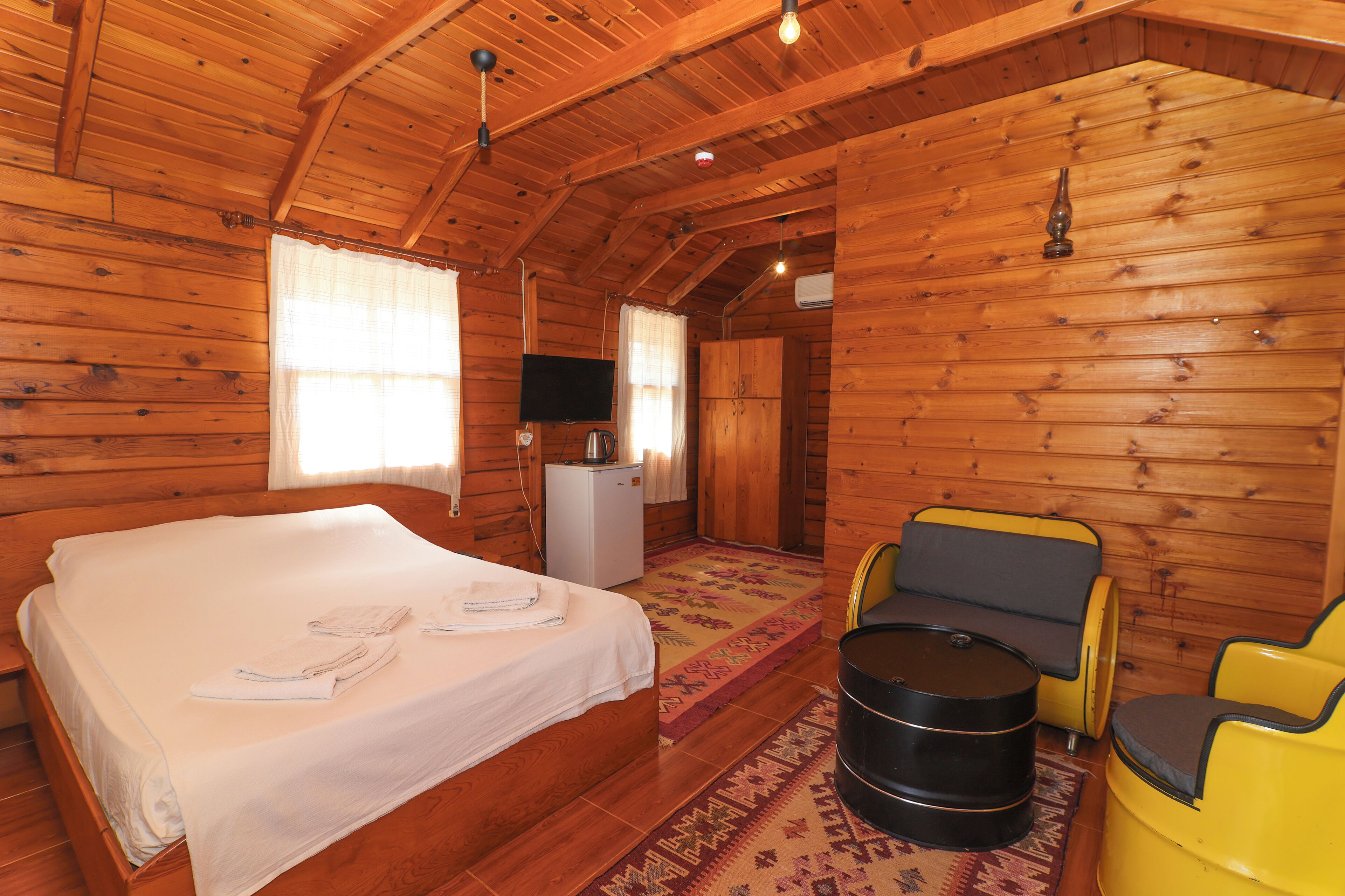 Quarto de cabana de madeira com uma cama, uma pequena cozinha compacta, uma televisão e um espaço de estar com uma mesa redonda e cadeiras.