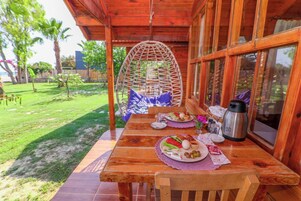 Una mesa de madera con platos de comida, un sillón colgante y una ventana con vistas a un jardín.