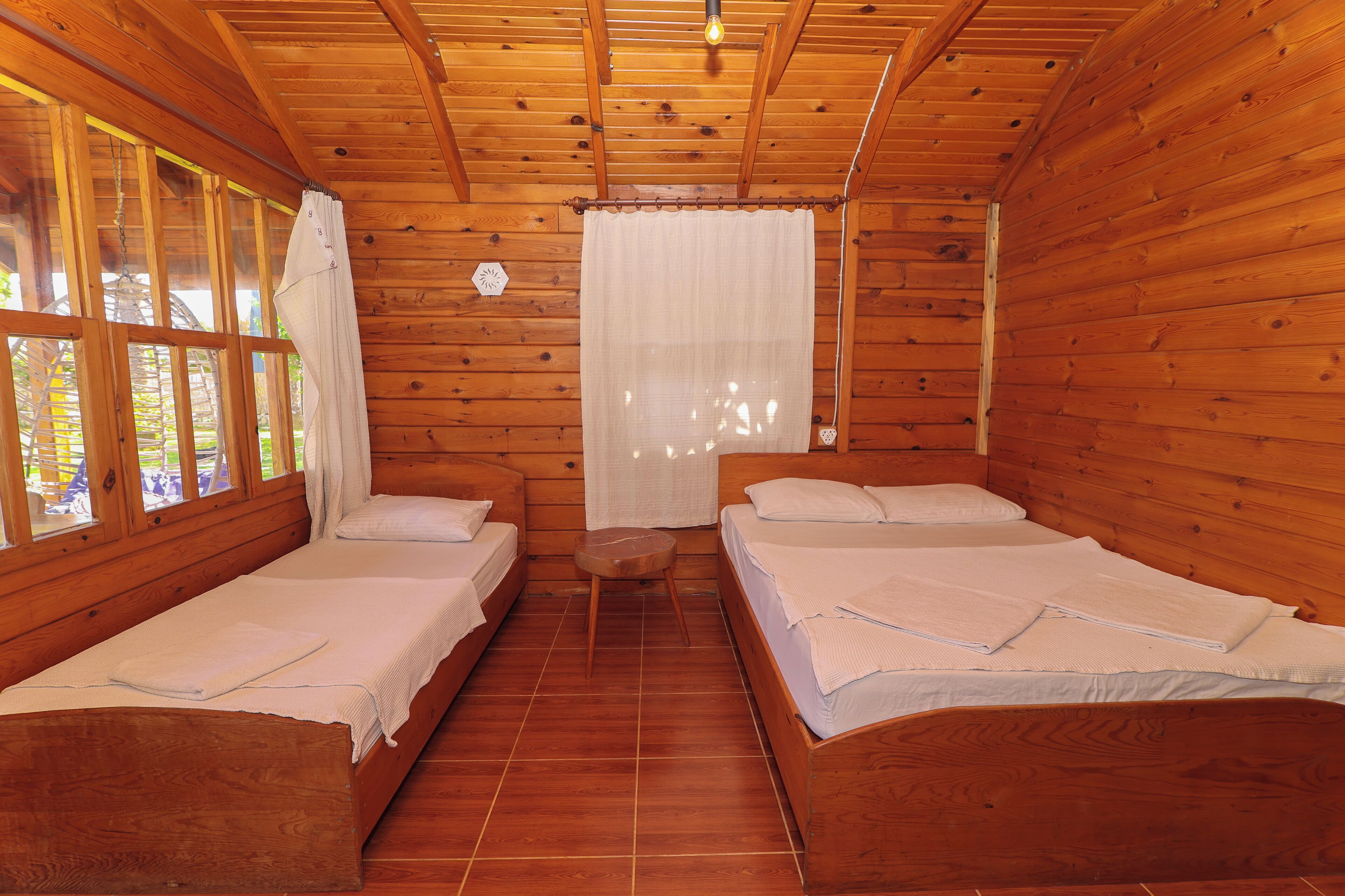Una cabaña de madera con dos camas individuales, un taburete pequeño de madera y una ventana con cortina.