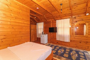 A wooden cabin room with a bed, a small white refrigerator, a television, and a patterned rug. - Demre Sahildeki Ev Bungalov (Demre)