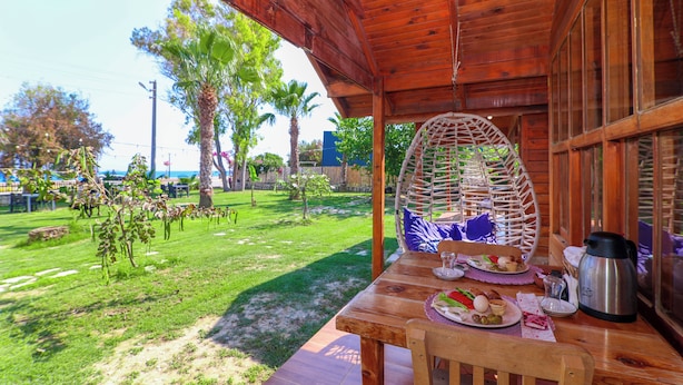 A wooden table with breakfast items, a wicker chair, and a view of a grassy area with trees. - Demre Sahildeki Ev Bungalov (Demre)