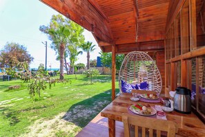 Una mesa de madera con alimentos para el desayuno, una silla de mimbre y vistas a un área verde con árboles.
