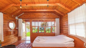 A wooden cabin room with a bed, a round mirror, a small table, and a window with a view of greenery. - Demre Sahildeki Ev Bungalov (Demre)