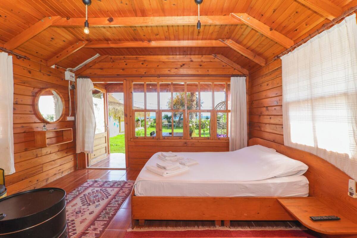 A wooden cabin room with a bed, a round mirror, a small table, and a window with a view of greenery.