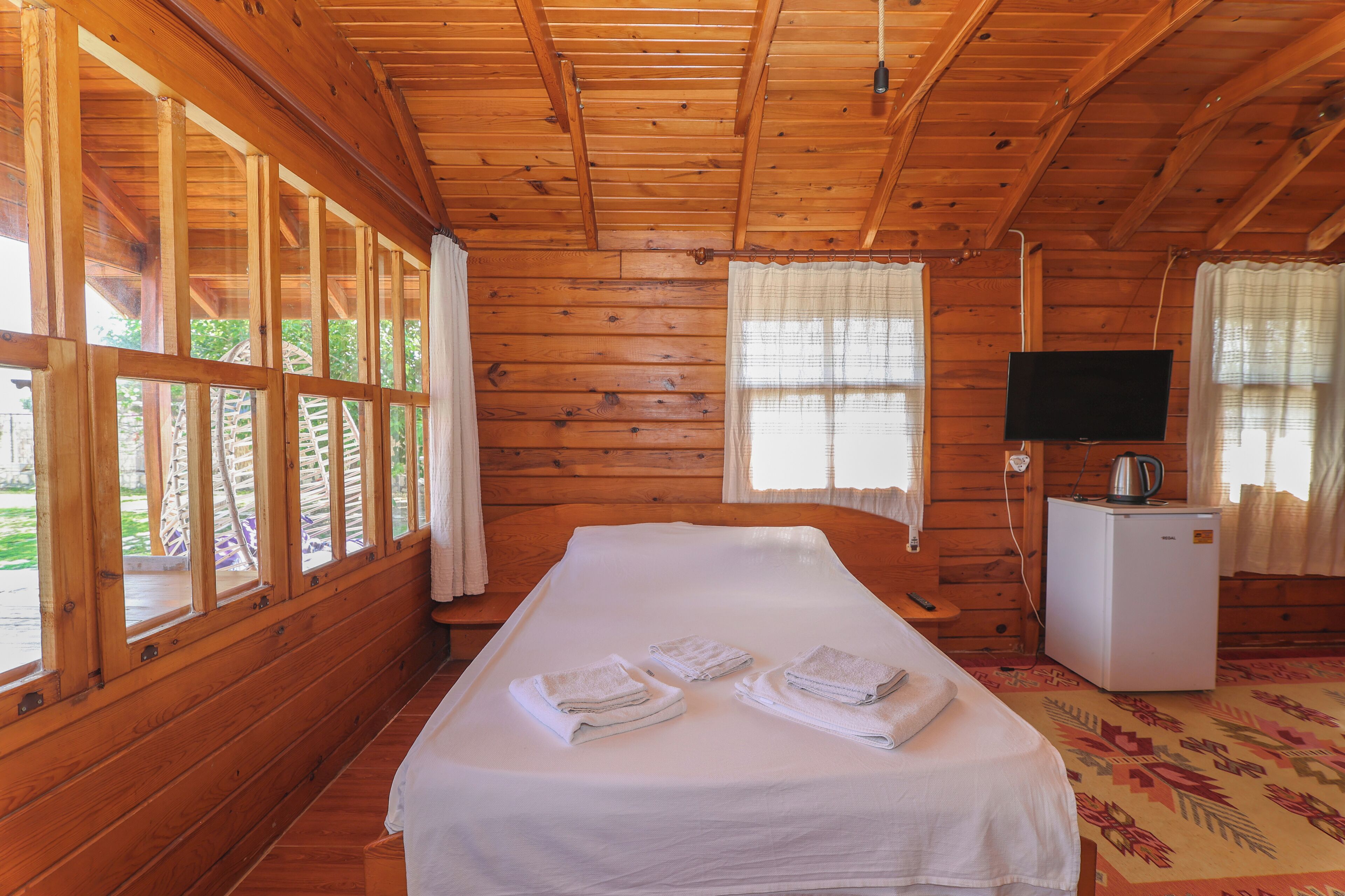 A wooden cabin room with a bed, towels, a television, and a refrigerator.