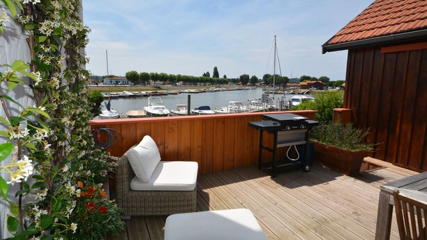 Eine Holzterrasse mit einer Korbusche und einem Grill, mit Blick auf einen Hafen mit Booten.