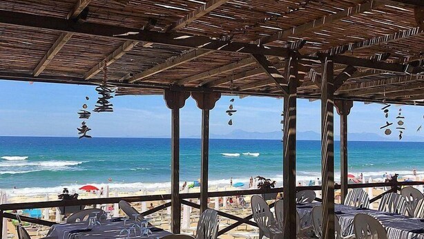 Un espace de restauration en plein air, avec des tables et des chaises, donnant sur une plage où l’on peut admirer les vagues et les parasols.