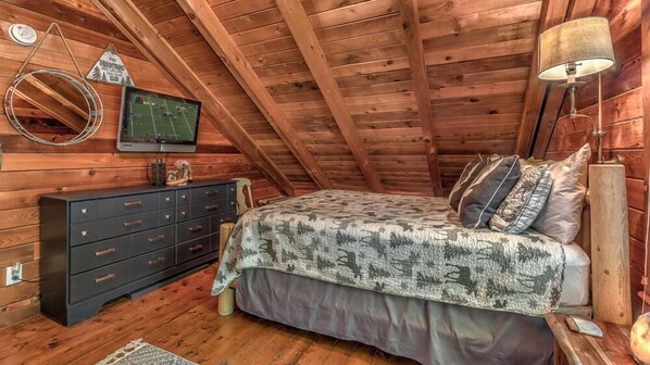 A cozy attic bedroom with a wooden ceiling, a bed with patterned bedding, a dresser, a television, and a lamp.
