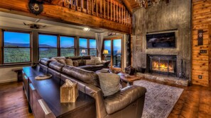 A spacious living room with a large leather sectional sofa, a fireplace, and large windows offering a view of the mountains.