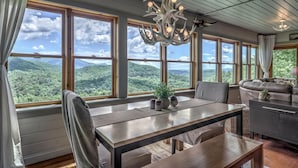 A dining area with a view of a mountainous landscape through large windows, a modern chandelier, and a leather sofa. - Top of the Ridge Chalet - Pet Friendly | Mountain Views | Hot Tub | Multiple Decks (Blue Ridge)