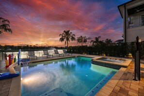 Una piscina con aro de baloncesto, sillas de descanso y un jacuzzi, rodeada de palmeras y vista al atardecer.
