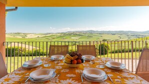 Mesa posta para quatro pessoas, com uma tigela de frutas no centro, com vista para uma paisagem campestre deslumbrante.