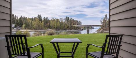 Un patio con dos sillas y una mesa, con vistas a un área verde, un puente y una masa de agua.