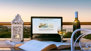 A laptop on a table with a webpage open, a wine bottle, a glass of wine, and a decorative lantern. - Romanza Villas (Sitia)