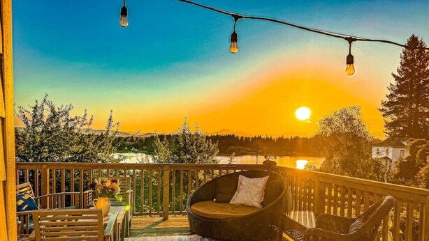 A wooden deck with outdoor furniture, including a sofa and chairs, overlooking a scenic landscape with trees and a body of water during sunset. - Sunset Garden Retreat-Sea & Mountain View w/ Sauna (Bremerton)