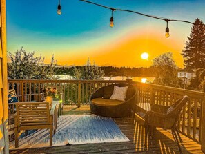 Una terraza de madera con mobiliario exterior, incluyendo un sofá y sillas, con vistas a un paisaje escénico con árboles y una masa de agua al atardecer.