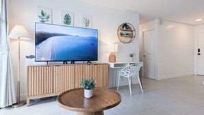 A modern living room with a large flat-screen TV, wooden paneling, a round wooden coffee table with a plant, a white chair, and a desk with a lamp. - High Floor 2BR/2BA+Den 5beds Partial Ocean View + 1 Parking 1 min from the beach (Honolulu)