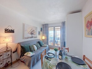 A cozy living room with a sofa, a dining table set for two, a balcony with a view, and a 'Bienvenue' sign.