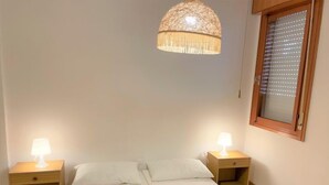 A bedroom with a bed, two bedside tables with lamps, a hanging light fixture, a window with blinds, and patterned floor tiles. - Canaima by Interhome (Bibione)