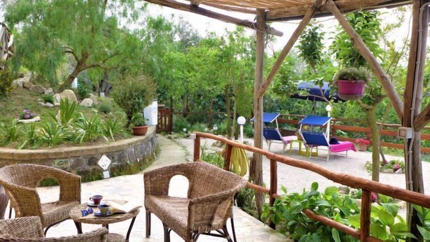 Area patio esterna con mobili in vimini, pavimento in pietra ed una vista su giardino con diverse piante e un pergolato in legno.