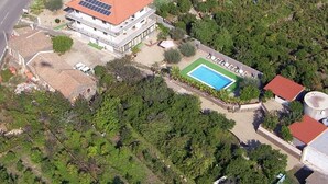 An aerial view of a property with a swimming pool, a large building with a red roof, and surrounding greenery.