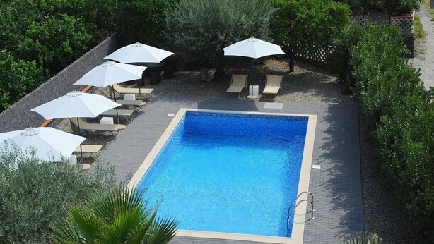 A square swimming pool with a tiled deck, surrounded by lounge chairs and umbrellas.