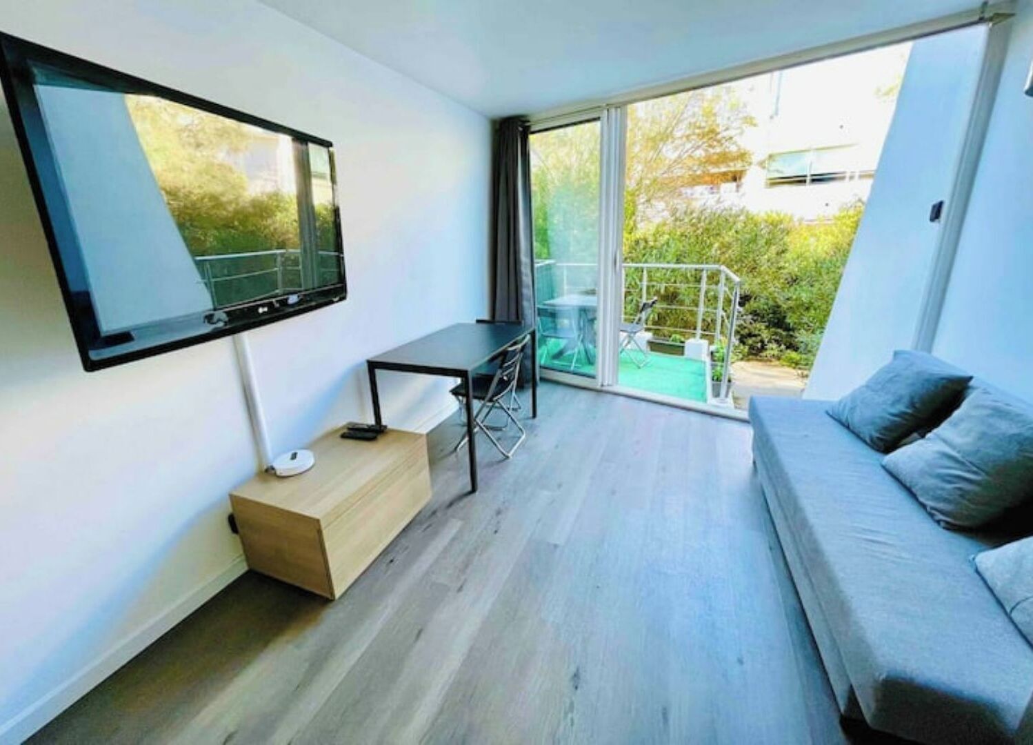 A modern living room with a flat-screen TV, a small table, a sofa, and a sliding glass door leading to a balcony with outdoor furniture.