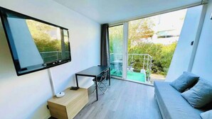 En moderne stue med et fladskærms-tv, et lille bord, en sofa og en skydedør, der fører ud til en balkon med havemøbler.