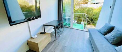 A modern living room with a flat-screen TV, a small table, a sofa, and a sliding glass door leading to a balcony with outdoor furniture.
