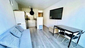 En moderne stue med en grå sofa, et spisebord med stole, et fjernsyn monteret på væggen og et køkken i baggrunden.