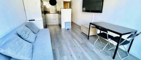 Een moderne woonkamer met een grijze bank, een eettafel met stoelen, een aan de muur bevestigde televisie en een keuken in de achtergrond.
