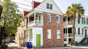 Une maison à deux étages, construite en briques, avec une porte verte, un balcon et un petit jardin devant.