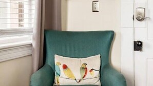 Un fauteuil de couleur turquoise avec un coussin à motif d’oiseau, un tableau accroché au mur et une fenêtre avec des stores.