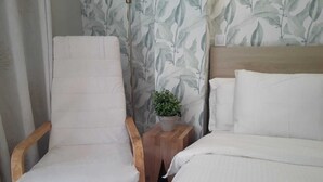 Ein Zimmer mit Holzstuhl, einem kleinen Nachttisch mit Topfpflanze und einem Bett mit weißer Bettwäsche.
