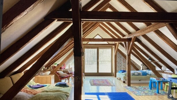 Une chambre mansardée avec un plafond en pente, des poutres en bois, un lit, un bureau et une fenêtre.