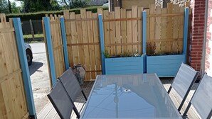 An outdoor patio with a glass table and four chairs, surrounded by a wooden fence and blue planters. - Charming house for 4 near town center (Sarzeau)