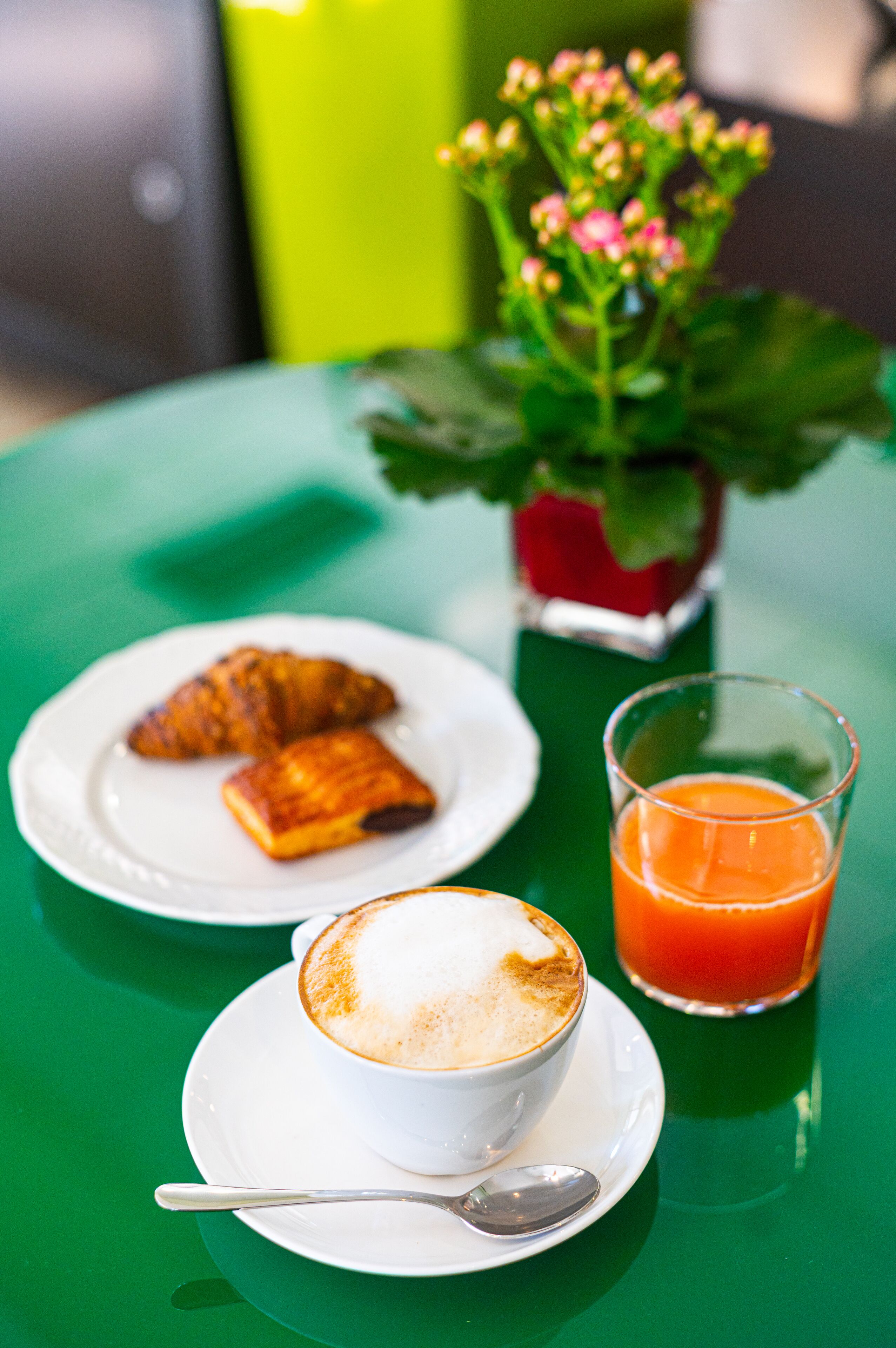 Une table recouverte d’une nappe verte, une assiette de croissants, une tasse de café, un verre de jus d’orange et une plante en pot.