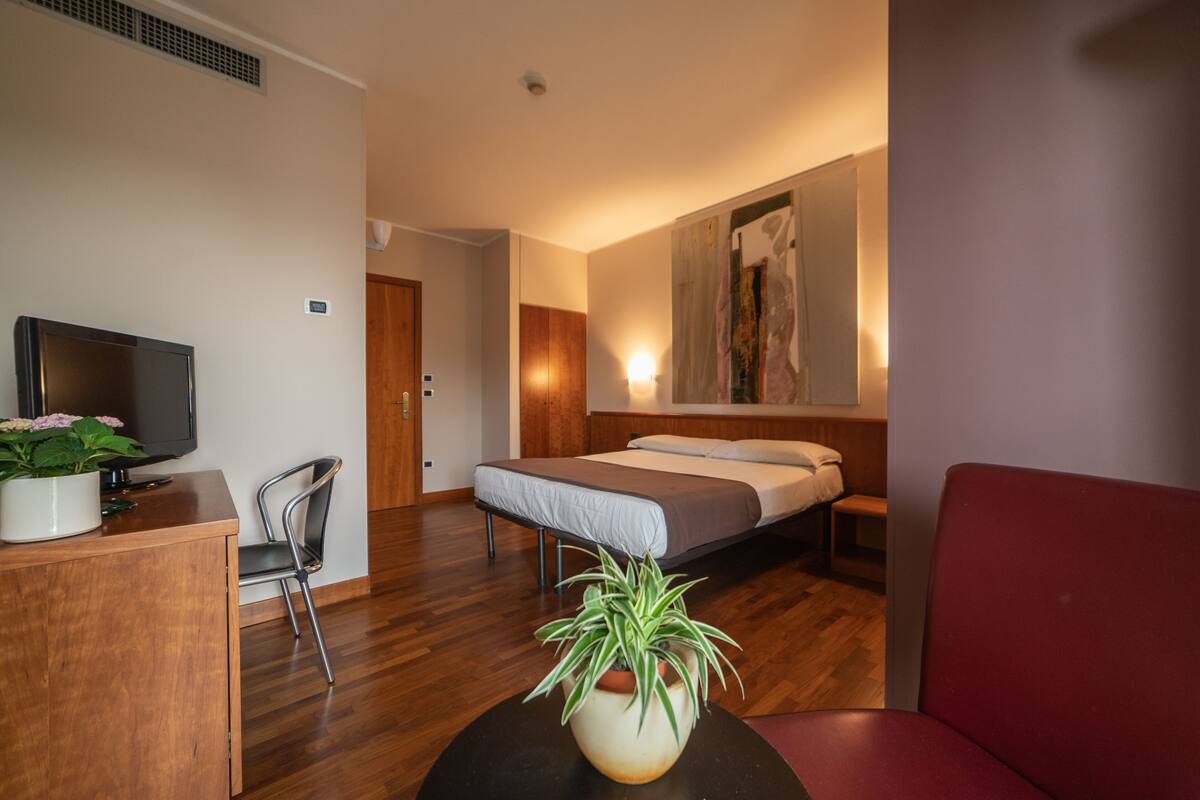 Ein Hotelzimmer mit Bett, Schreibtisch mit Fernseher, Stuhl, rotem Sofa und einer Zimmerpflanze.