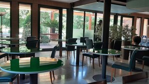 Een moderne, overdekte eetruimte met ronde tafels en stoelen, grote ramen en potplanten.
