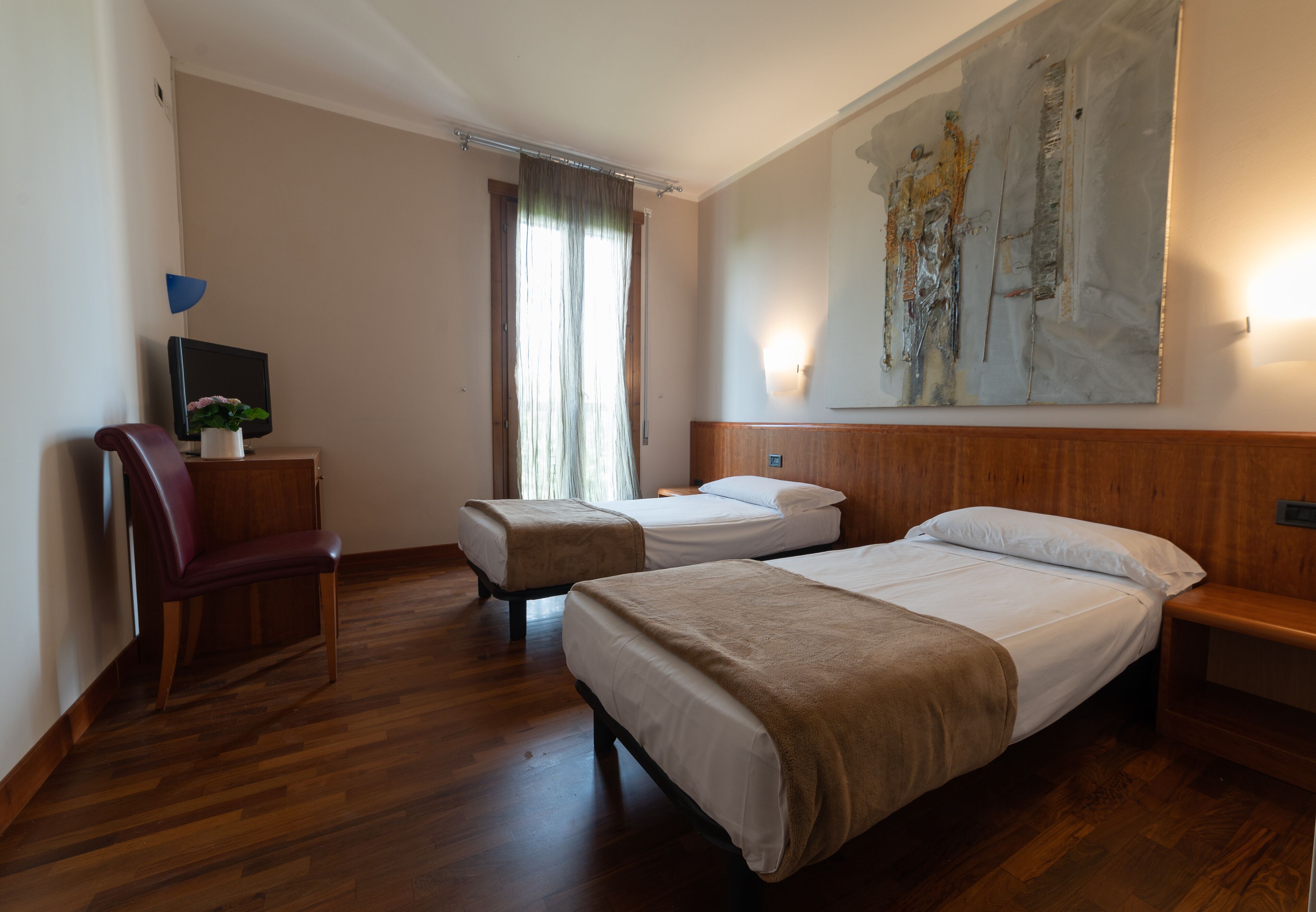 A hotel room with two beds, a wooden floor, a desk with a chair, a painting on the wall, and a window with curtains.