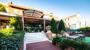 Een hotelentree met een overdekte gang, trappen naar de ingang en een buitenzitplaats met tafels en stoelen.