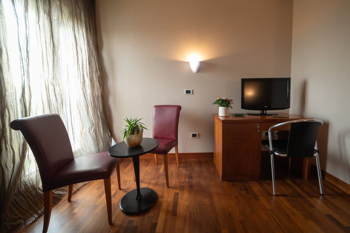 Ein Zimmer mit Holzboden, einem Fernseher auf einer Holzkommode, einer Topfpflanze, einem roten Sessel, einem schwarzen Stuhl und einem Tisch.