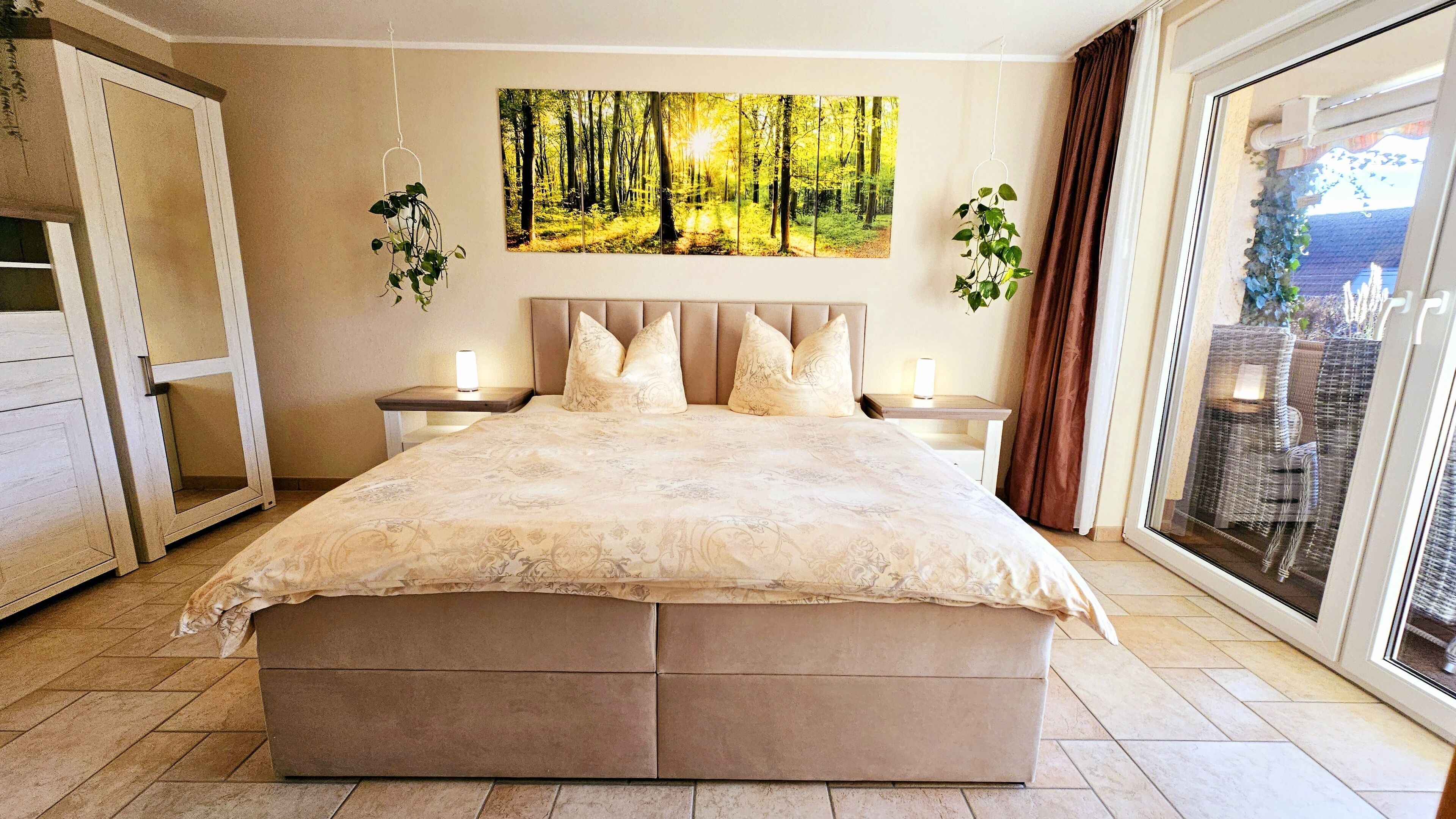 A bedroom with a large bed, two bedside tables with lamps, a painting of a forest, and a view of outdoor furniture through a sliding door.