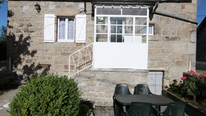 Un edificio de piedra con un pequeño patio amueblado con una mesa y sillas, una ventana con contraventanas y una puerta.